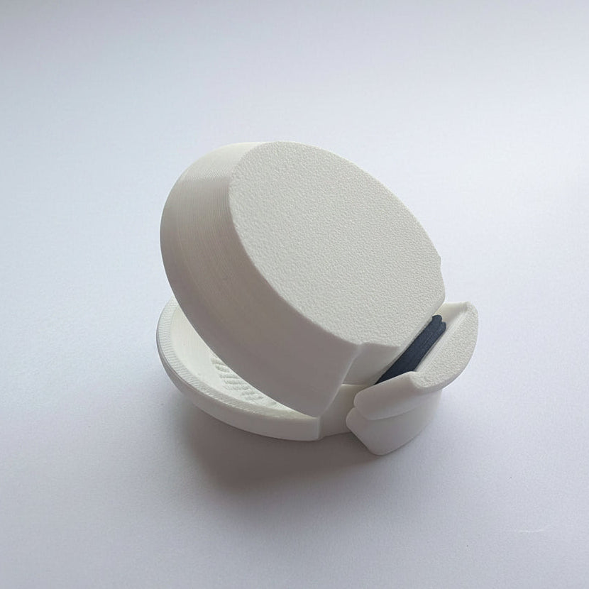 A white 3D-printed castanet, shown open on a white surface with other colorful items blurred in the background.